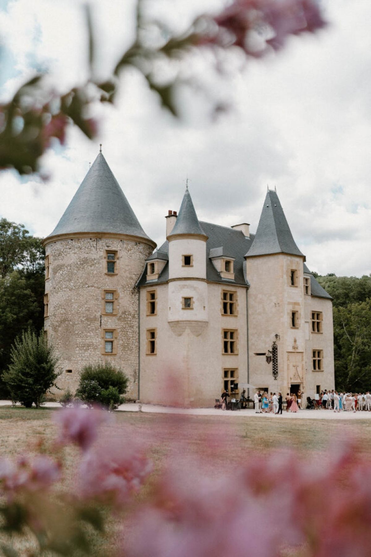 Limestone chateau with conical slate towers and cocktail hour guests framed by pink flowering branches