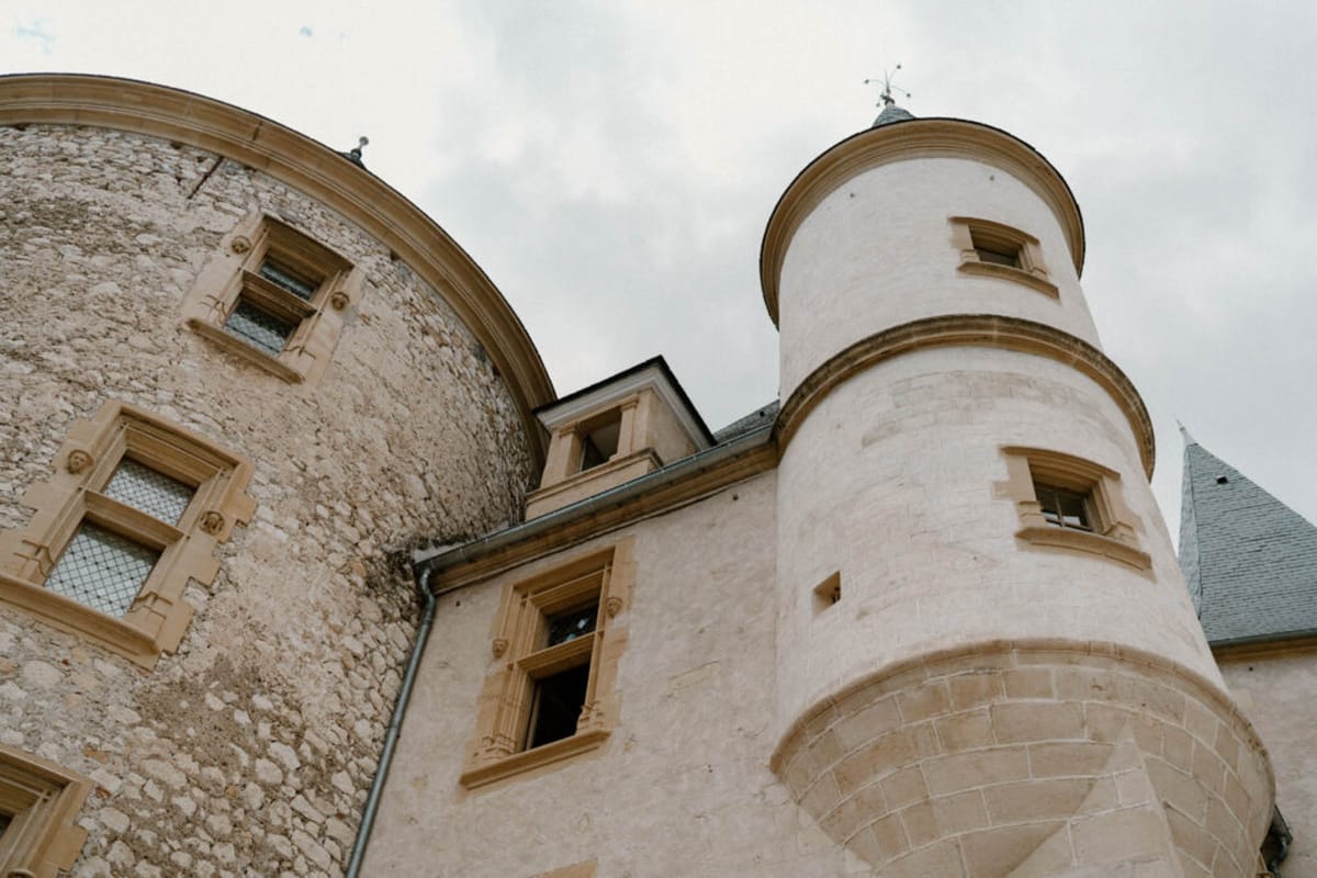 An upward-angled architectural detail shot of a French château, showing two distinct tower structures: a large rounded medieval stone tower on the left with rough-cut pale limestone masonry and diamond-paned windows with warm sandstone surrounds, and a smoother rendered circular turret on the right with slate-capped conical roof and stone-framed windows. A third slate roof is partially visible at the far right. No people are present in the frame. The composition is a low-angle wide shot looking up toward an overcast sky, emphasizing the height and architectural character of the building. Potential venue feature image.