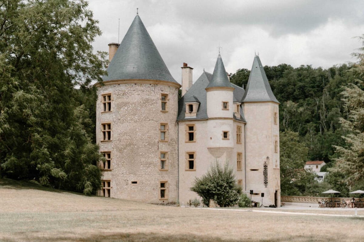 A wide exterior shot of a French château featuring multiple round towers with pointed slate-grey conical roofs and cream-coloured limestone facades with warm stone window surrounds. The architecture is classic French Renaissance style, with four visible turrets of varying heights forming the main structure. To the right side of the building, a terrace area is visible with white parasols and what appears to be a small group of guests gathered, along with audio equipment suggesting an outdoor event in progress. The shot is taken from across an open lawn, framing the full building against a backdrop of dense woodland. Potential venue feature image.