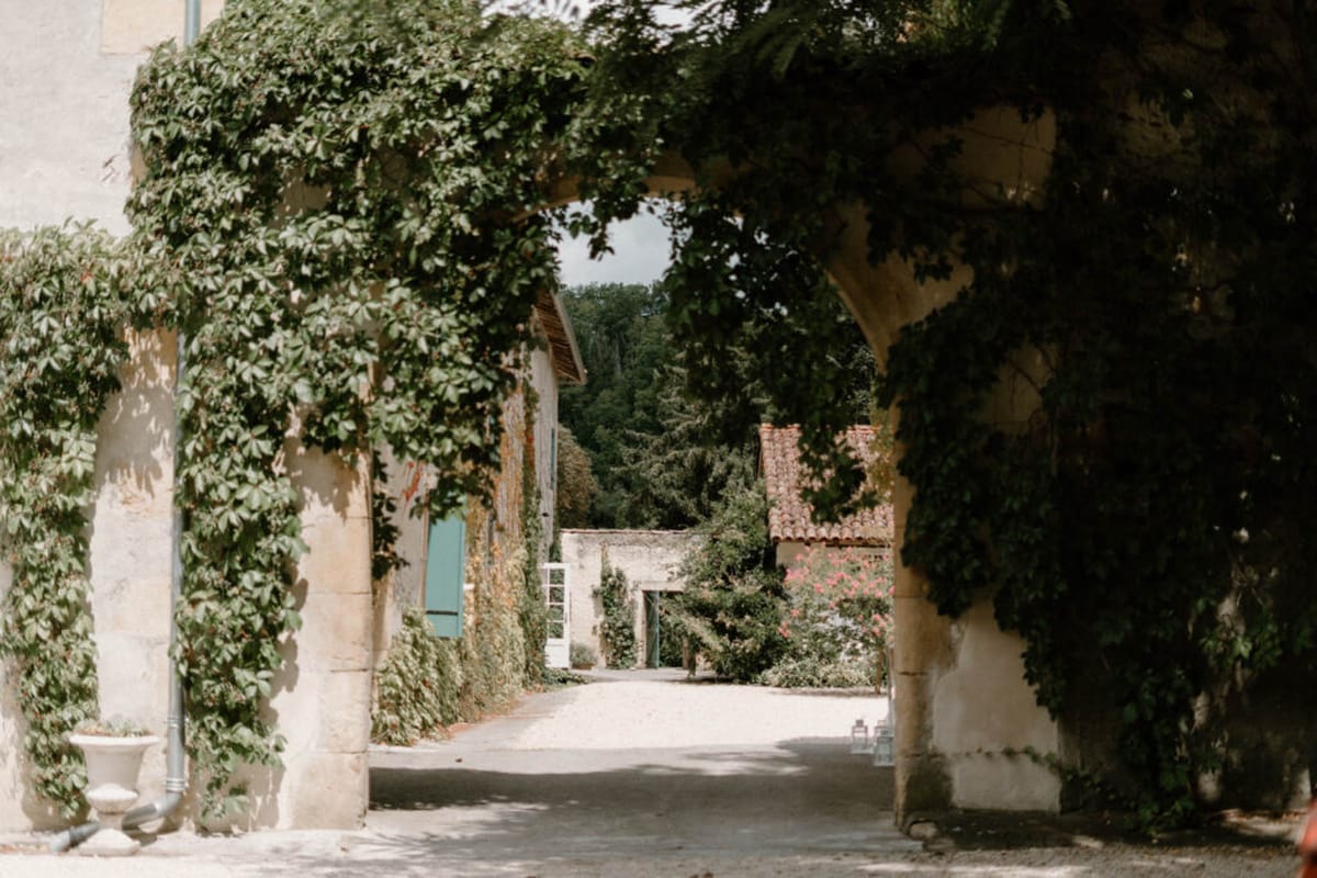 A wide shot of a French country estate courtyard viewed through an archway framed by ivy-covered stone pillars. The scene shows a gravel pathway leading between cream-colored rendered stone buildings with teal-painted shutters and terracotta-tiled roofs, all heavily draped in climbing green vines. A white urn planter is visible at the base of the left pillar, and pink climbing roses can be seen on a building in the middle distance. No people are present in the image. Potential venue feature image.