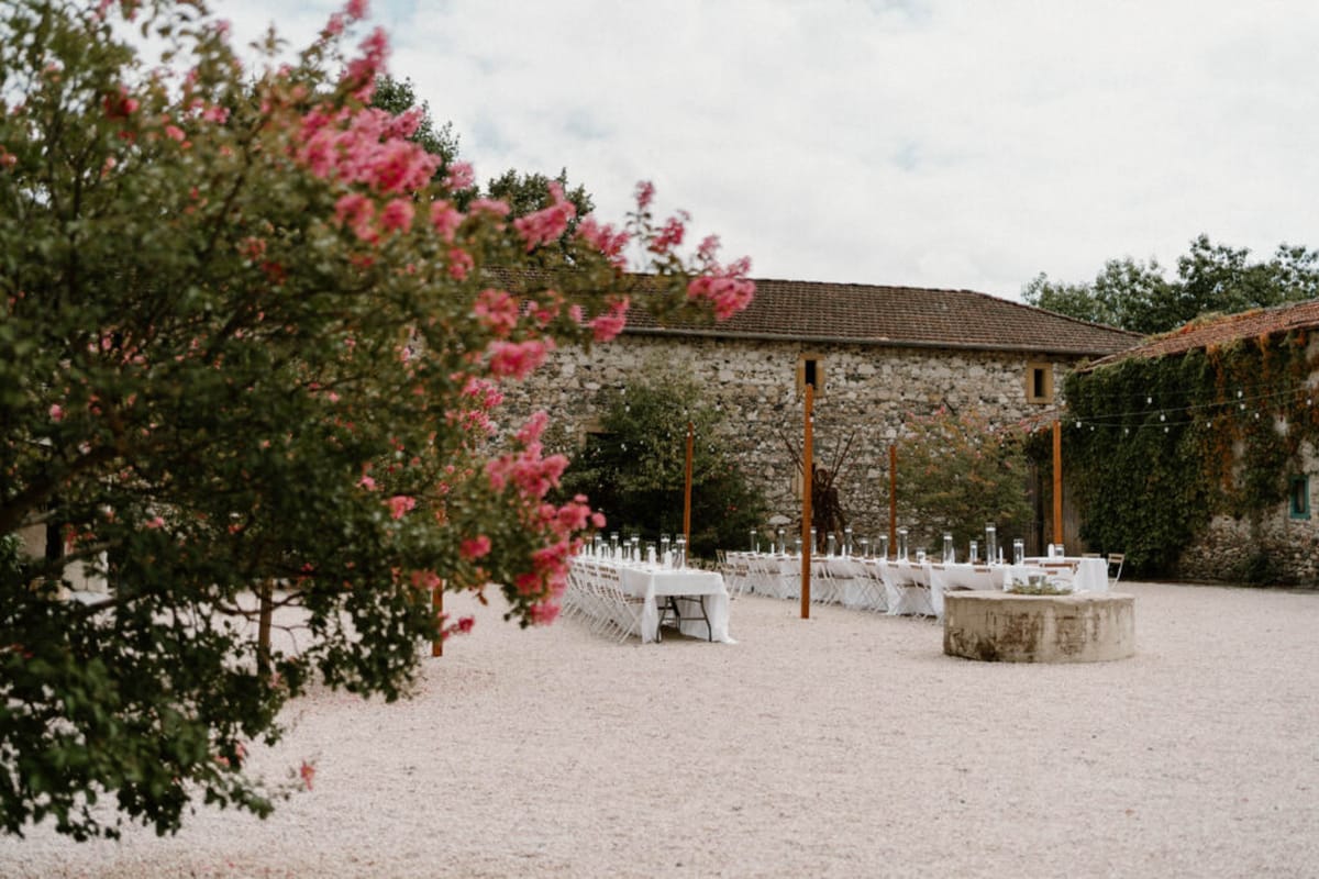 An outdoor wedding reception setup in the courtyard of a French stone farmhouse or domaine, photographed as a wide shot with a blossoming pink crêpe myrtle tree in the foreground. Two long banquet tables are dressed in white linen and lined with white folding chairs, set on a gravel courtyard. Each table is decorated with a row of tall, slender glass candleholders or bud vases running down the center. Wooden upright posts are positioned between the tables, strung with café-style bistro lights. A low stone basin or well feature sits to the right of the tables. The decor palette is minimal and clean — white linens, clear glass, natural wood — suggesting a rustic-modern styling approach. Potential venue feature image.
