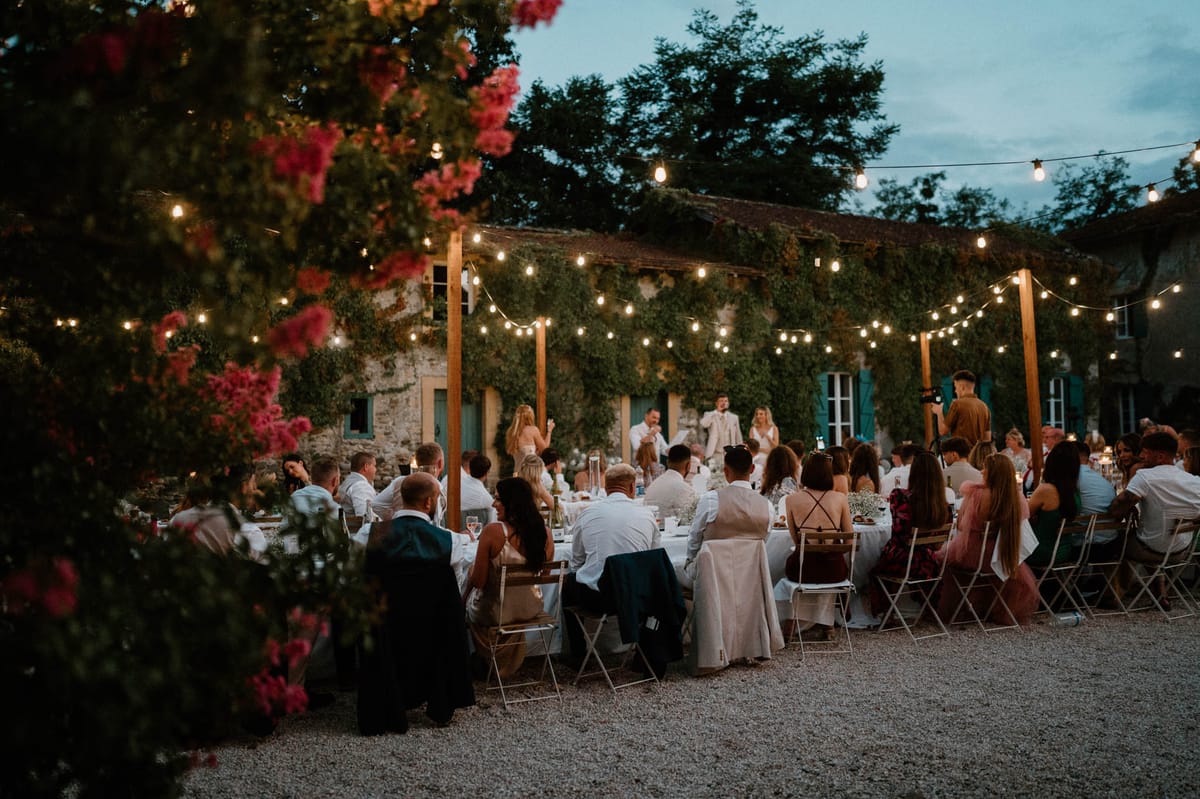 An outdoor wedding reception dinner is underway at dusk, with approximately 40–50 guests seated along long banquet-style tables set on a gravel courtyard. A speech is being delivered at the head of the table, where the bride in a white gown and groom in a light beige suit are seated alongside a standing speaker. The venue is a ivy-covered stone farmhouse with teal-painted shutters and doors, typical of French rural architecture. Warm-toned festoon bulb lights are strung across wooden posts above the tables, providing the primary lighting as daylight fades. Table linens are white, with tall glass lanterns and small floral arrangements visible as centerpieces. Guests are dressed in a mix of formal and semi-formal attire including blush, dusty rose, burgundy, and white outfits. A flowering shrub with deep pink blooms is partially visible in the foreground, framing the left side of the wide-angle shot. Potential venue feature image.