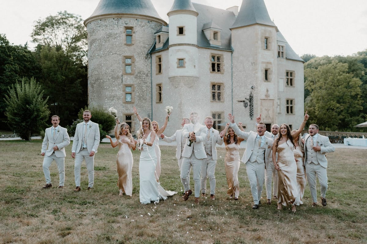 Wedding party walks toward camera as groom pops champagne in front of twin-towered French chateau