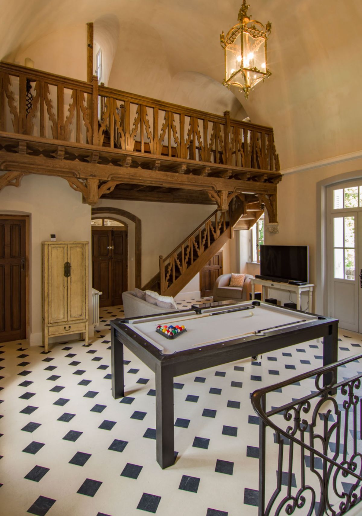 Interior wide shot of a grand room inside what appears to be a French château or manor house, featuring no wedding-related activity or people. The room includes a black-framed pool table with a grey felt surface and racked billiard balls in the foreground, set on a black-and-white diamond-patterned tile floor. A carved dark wood staircase with ornate gothic-style balustrade leads to an upper mezzanine level with matching carved railings. A brass and glass lantern pendant light hangs from the vaulted ceiling. Additional furnishings include a distressed cream painted armoire, a grey sofa, and a flatscreen television on a white console. A wrought iron stair railing is partially visible at lower right. Potential venue feature image.