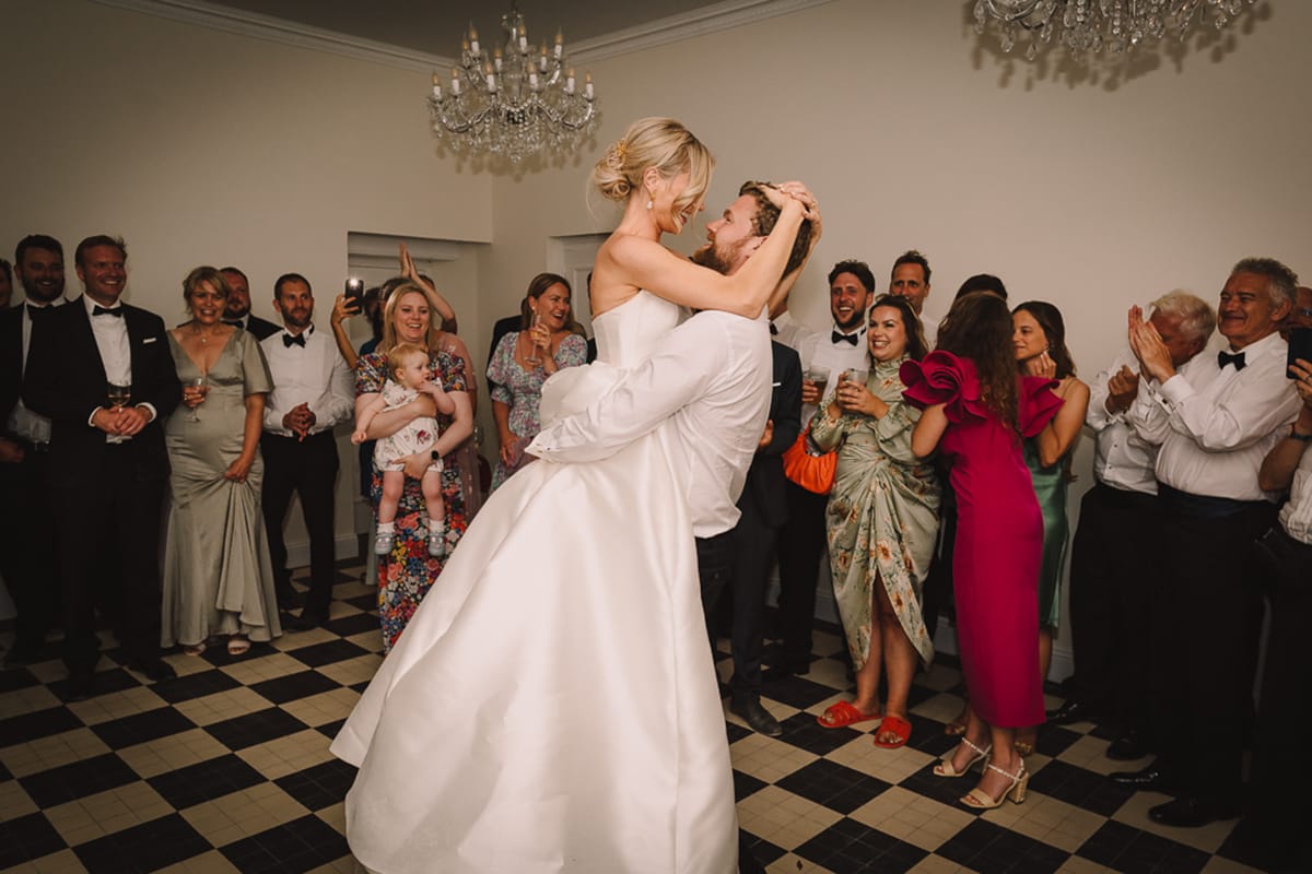 The first dance is taking place in an indoor ballroom-style reception space, where the groom — wearing a white dress shirt with the sleeves rolled up — has lifted the bride off the ground as they share a close, joyful moment face-to-face. The bride wears a strapless ivory satin ballgown with a structured bodice and a full skirt, with her blonde hair in an updo accessorized with a gold hair piece. Approximately 20 guests form a circle around them on a black-and-white checkered tile floor, laughing, clapping, and reacting enthusiastically; guests are dressed in formal attire including black tuxedos, a fuchsia pink ruffle-shoulder midi dress, a sage green satin gown, and a floral maxi dress. The room features white walls with crown molding and a crystal chandelier overhead, creating a classic, formal interior setting. The shot is a wide, documentary-style action portrait capturing the couple in the foreground with the crowd visible in the background.