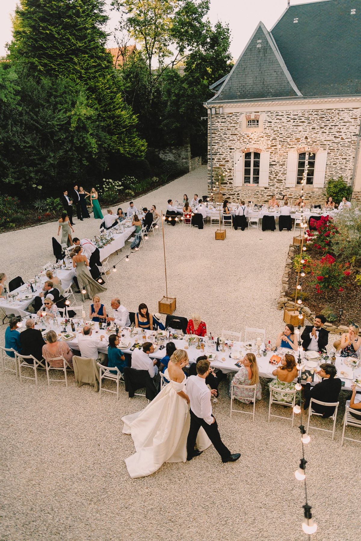 An outdoor wedding reception dinner is underway on a gravel courtyard in front of a traditional French stone manor house with a slate roof. The couple — the bride in a white strapless ballgown with a full skirt and the groom in a black tuxedo and white shirt — walk together through the center of the seating arrangement, likely making their entrance or greeting guests. Long white-linen banquet tables are arranged in a curved U-shape around a central open space, with approximately 60–80 guests seated in white folding chairs and mismatched occasional chairs. The tables are set with glassware, wine bottles, candles, and low floral centerpieces. Edison bulb string lights are strung on wooden pole stands along the tables, adding warm ambient lighting as the evening sets in. Guests are dressed in a mix of formal attire including teal, emerald green, navy, and patterned dresses. The overall decor palette is relaxed and classic, with white linens and warm candlelight creating a convivial atmosphere. Wide aerial shot taken from an elevated angle above and behind the couple.