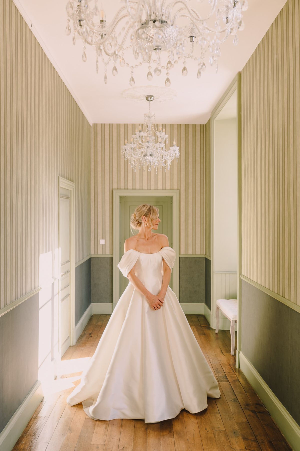 Bride in ivory off-shoulder ballgown with draped sleeves in hallway with sage striped wallpaper and chandeliers