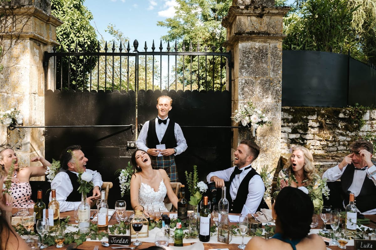 Kilted speaker drawing laughter from head table with greenery runner white roses bride and groom before stone gate