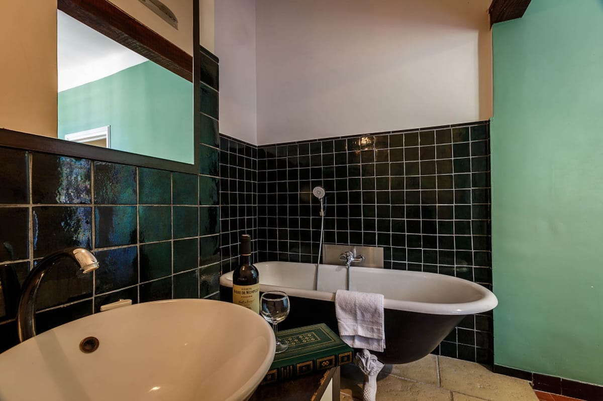 This image is not a wedding photograph but rather an interior shot of a bathroom within what appears to be a venue property. The room features a black clawfoot freestanding bathtub with a white interior, set against dark teal-green square glazed tiles that cover the lower half of the walls. A white vessel sink with a black faucet is visible in the foreground. A bottle of red wine and an empty wine glass are staged on a small ornate green side table beside the tub, along with a folded white towel. The right wall is painted mint green, and a large mirror reflects the space. The floor appears to be stone tile. The styling is eclectic, mixing vintage and classic elements. Wide interior shot. Potential venue feature image.
