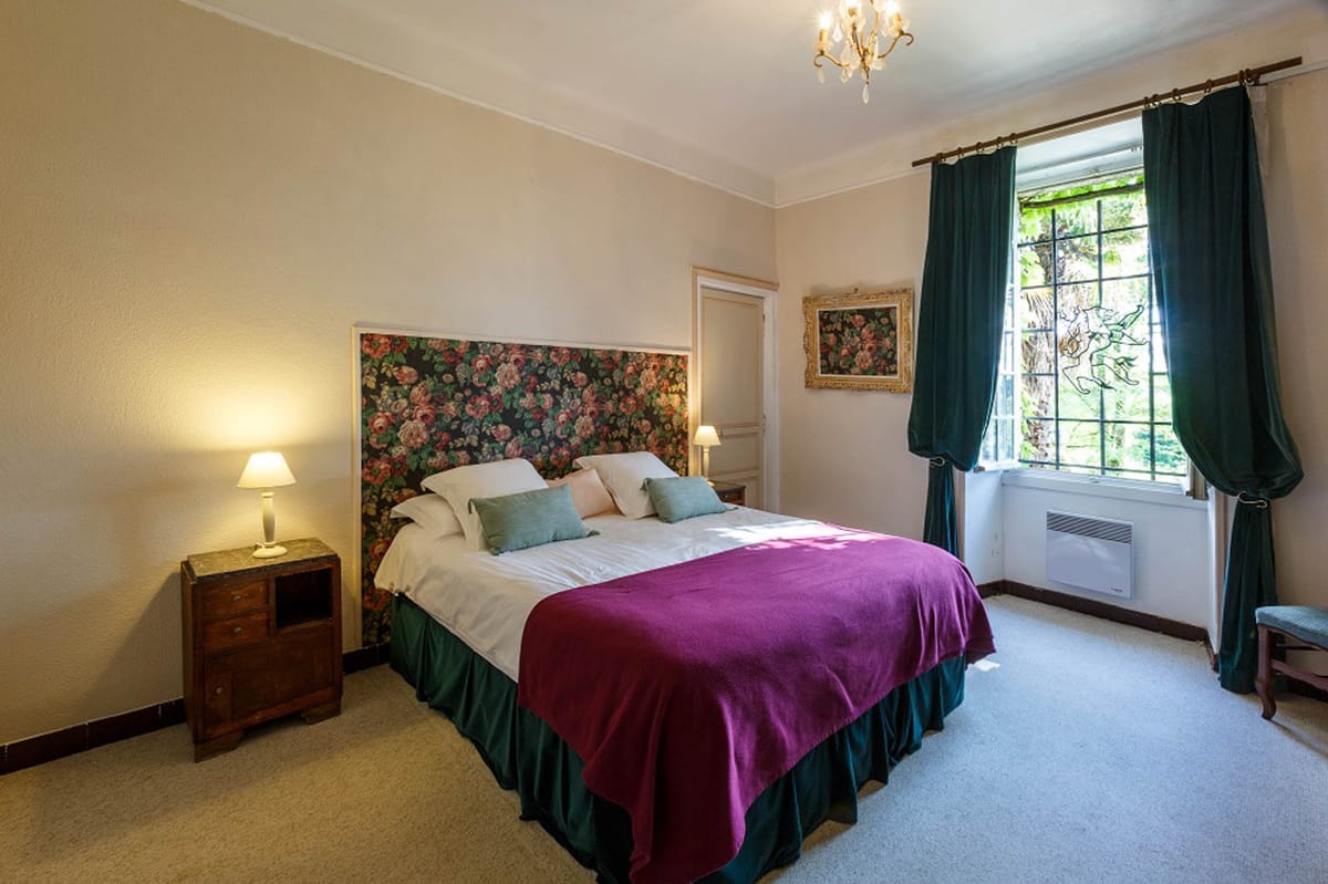 Wide-angle interior shot of a guest bedroom, likely within a château or manor house used as a wedding venue. The room features a double bed dressed in white linens with a magenta throw blanket and a dark green bed skirt, topped with sage green and blush accent cushions. The upholstered headboard is covered in a bold floral fabric with pink, coral, and green tones, framed by a cream border. A small gold chandelier hangs from the ceiling, and warm-toned bedside lamps flank the bed on wooden nightstands. Deep teal curtains frame a large iron-grid window overlooking greenery, and a gold-framed floral painting hangs on the adjacent wall. The decor style mixes classic French country elements with bold color choices. Potential venue feature image.