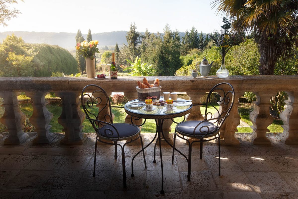 A wide shot of a stone balustrade terrace set for an intimate breakfast for two, with no people present. The terrace features a small round wrought-iron bistro table with a glass top and two matching wrought-iron chairs with dark navy cushions. The table is laid with a wicker basket of croissants, ceramic mugs, jam jars, and small condiments. On the stone balustrade ledge behind the table sit a champagne bottle, a vase of yellow and orange roses, a silver teapot, a small dish of fruit, and a glass vase. The setting has a classic French country styling with warm morning light casting long shadows across the stone-tiled floor. Potential venue feature image.