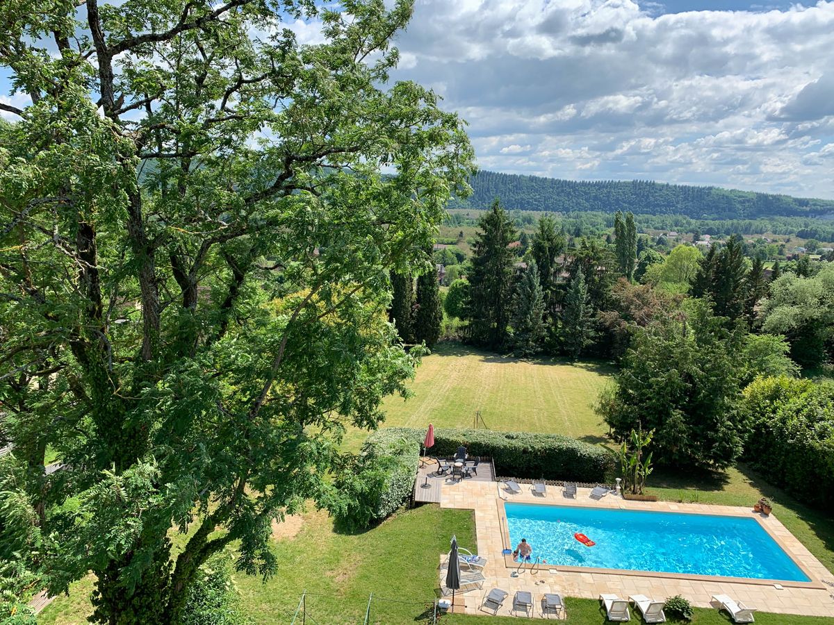 An elevated wide shot looking down over the outdoor grounds of a French country property, showing a rectangular swimming pool with bright blue water surrounded by a beige stone terrace with grey sun loungers. A wood-decked seating area with dark outdoor furniture and a closed red parasol sits adjacent to the pool, bordered by a clipped hedge. One person is visible standing at the pool's edge. Beyond the pool, a large open lawn extends toward tall cypress trees, with a broad valley landscape and forested hillside visible in the distance. No wedding activity or decor is visible in this image. Potential venue feature image.