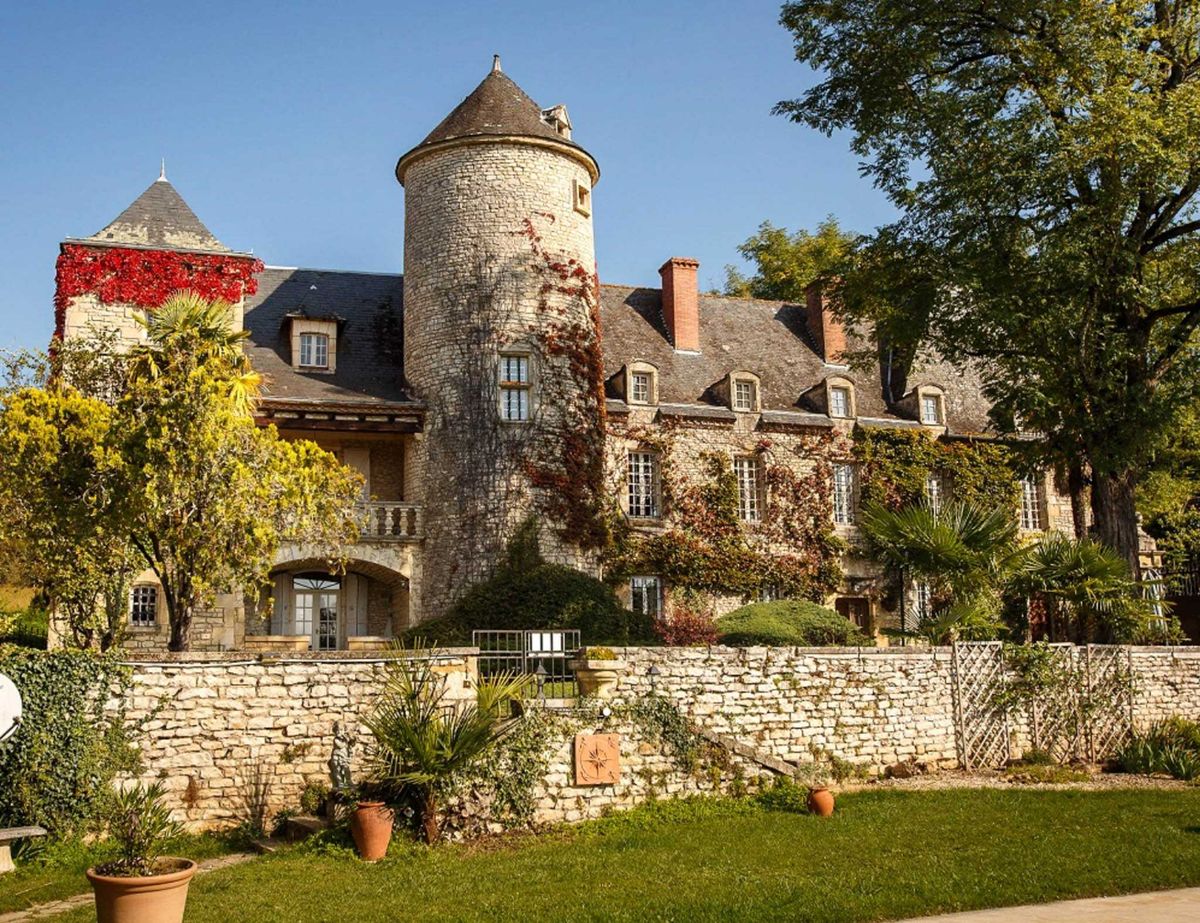 A wide exterior shot of a French château featuring a prominent round stone tower with a conical slate roof, multi-story limestone façade with dormer windows, and a slate mansard-style roof. The building is partially covered in climbing vines displaying deep red and burgundy autumn foliage, with additional red virginia creeper covering a second tower on the left. A low stone perimeter wall runs across the foreground, decorated with terracotta planters and a decorative medallion plaque. The grounds include a manicured lawn, palm trees, and mature deciduous trees in autumn yellow and green tones. No people are visible in the frame. Potential venue feature image.