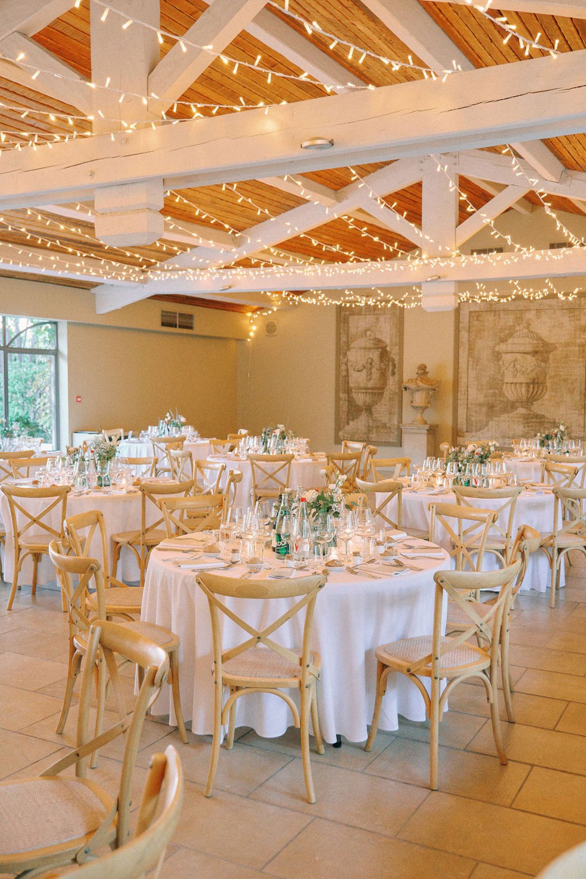 Indoor barn reception with fairy-lit ceiling, round tables, cross-back chairs, and classical fresco murals