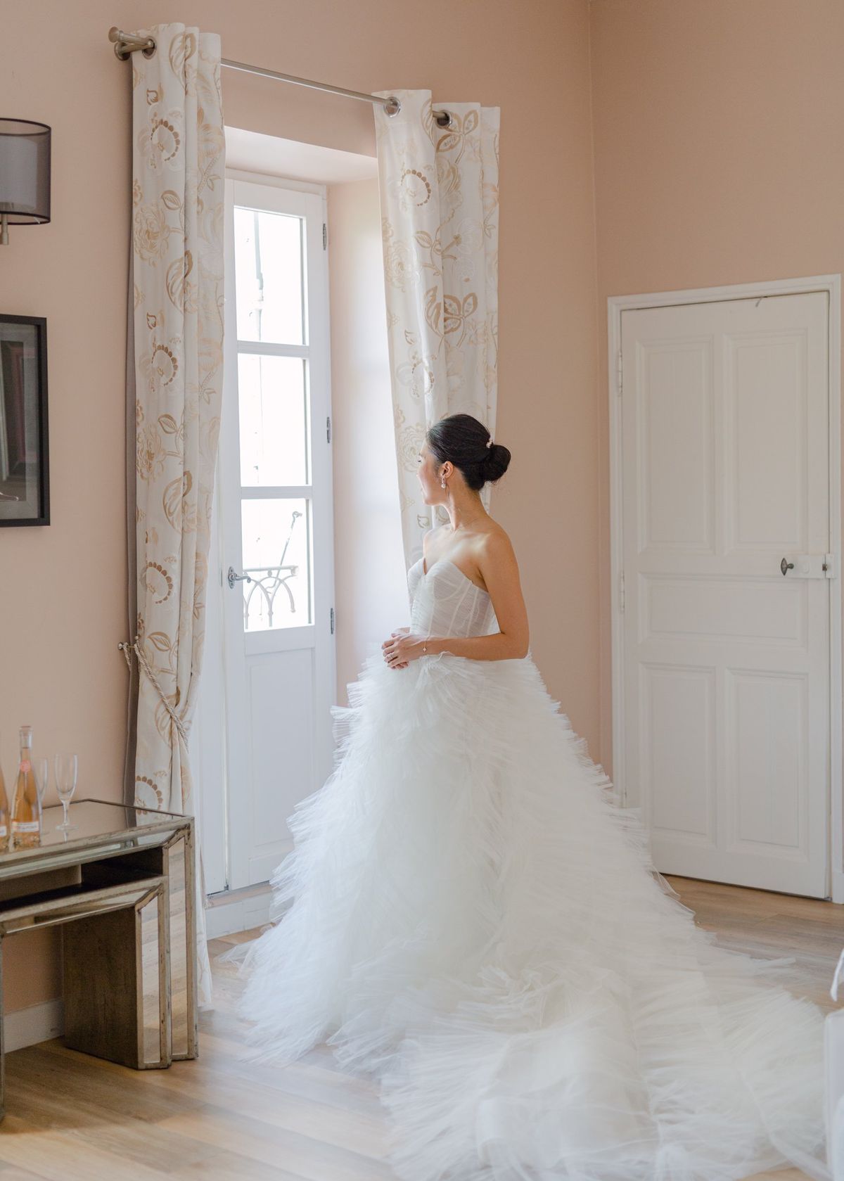 Bride in ruffled tulle ballgown gazing through French door in blush-walled room with rose and flutes