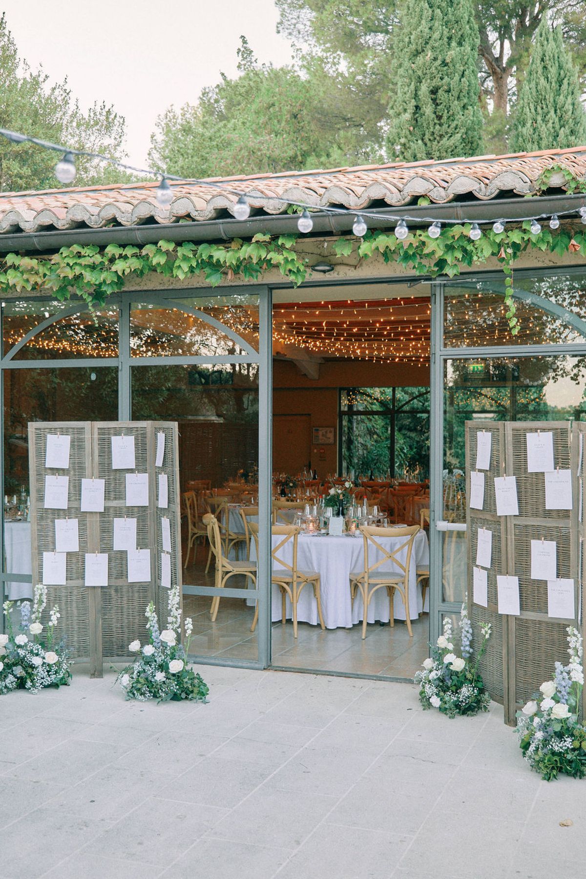 Glass pavilion reception entrance with wicker seating chart displays, blue and white flowers, fairy lights inside