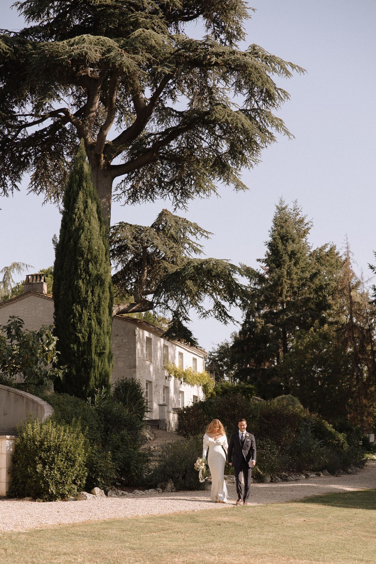 Bride and groom walking hand-in-hand across lawn of French country estate with manor house in background