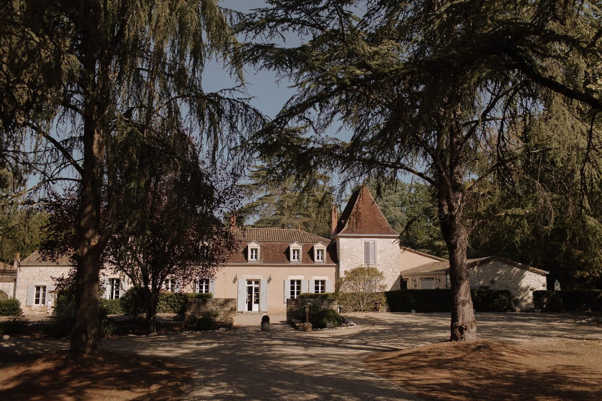 A wide exterior shot of a French country estate, showing a multi-building property with warm sand-toned rendered walls, white-framed windows fitted with pale blue-grey shutters, and terracotta tile rooflines. A pointed stone tower with a steep conical roof is visible behind the main house, indicating a historic manor or small château. The grounds include a gravel courtyard in the foreground, neatly trimmed hedging along the façade, and potted topiaries flanking the main entrance door. No people or wedding-specific decor are visible in the frame. The photograph is taken in natural daylight with a wide composition framing the buildings through mature trees. Potential venue feature image.