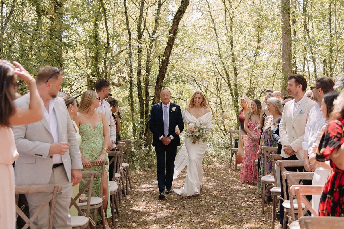 Bride in draped cape gown walked down woodland aisle with cross-back chairs by father in navy suit