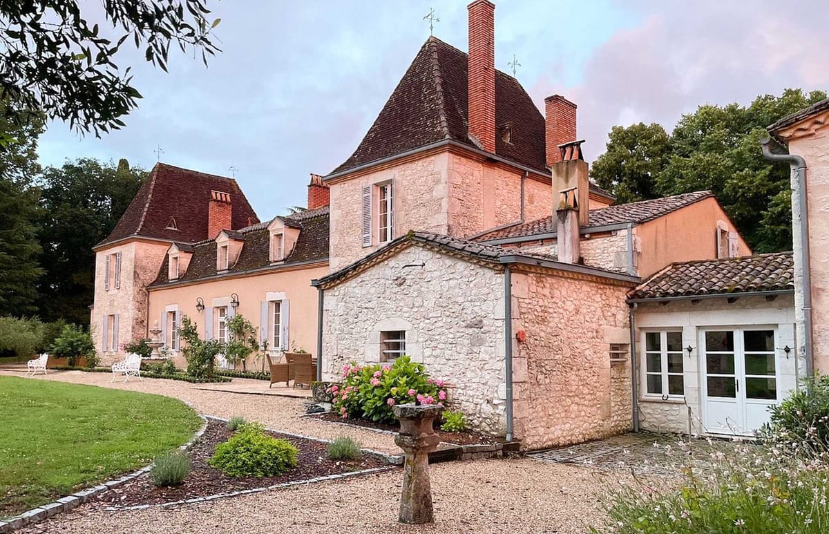 An outdoor wide shot of a French château and its outbuildings, featuring warm terracotta-pink rendered façades, dark tile rooftops with pointed turrets, multiple chimneys, and pale grey-blue shuttered windows. The grounds include a gravel courtyard with a stone pedestal planter holding pink and white hydrangeas, ornate white metal garden benches on the lawn, and a small stone fountain. A covered terrace area with wicker seating is visible along the main building frontage. No people are present in the image. Potential venue feature image.