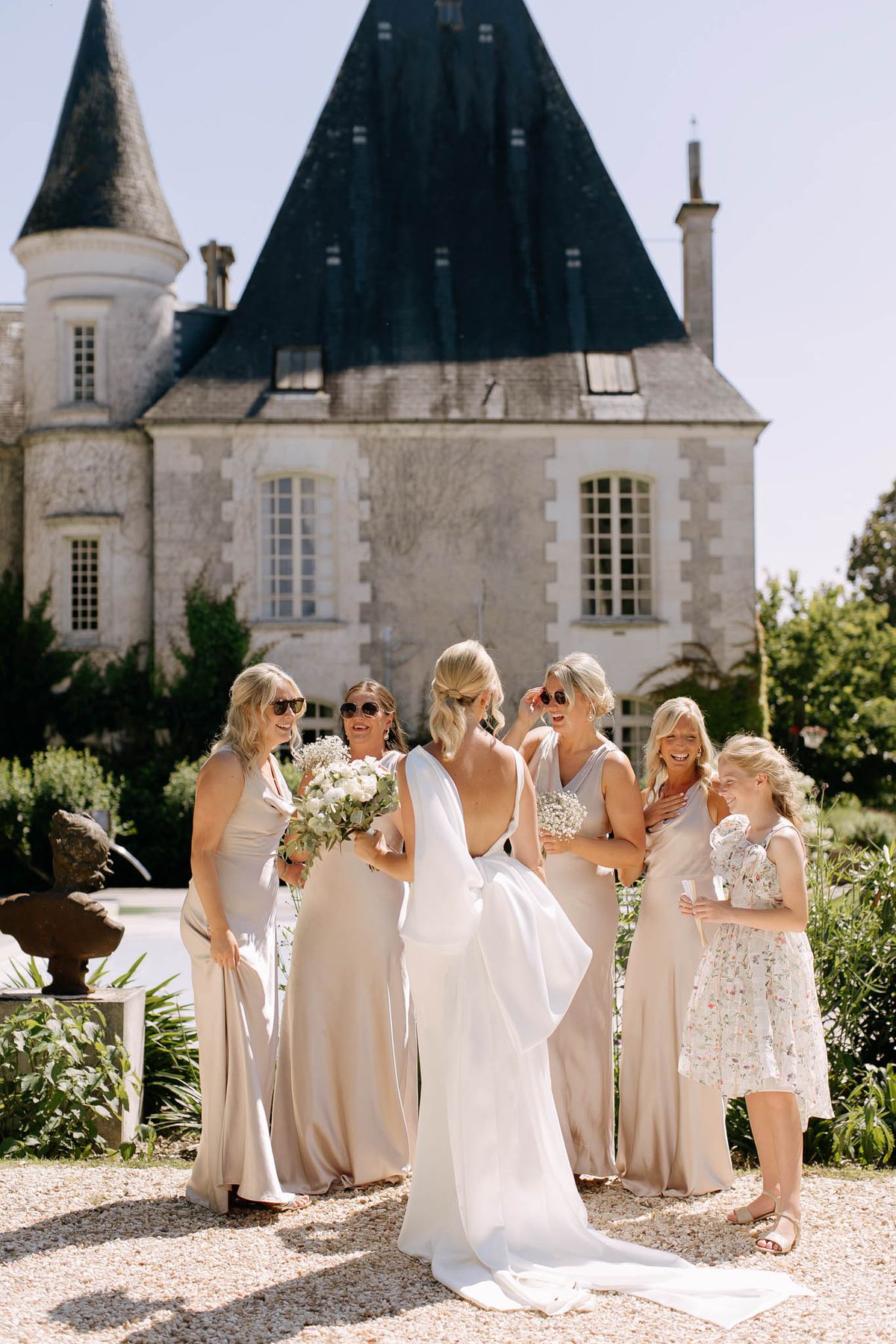 Bride with four bridesmaids in champagne satin dresses laughing on gravel courtyard before French chateau
