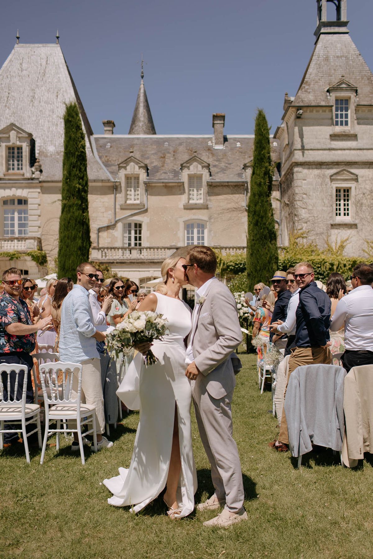 Couple kissing down aisle past 50 guests on white chairs before multi-turreted limestone chateau