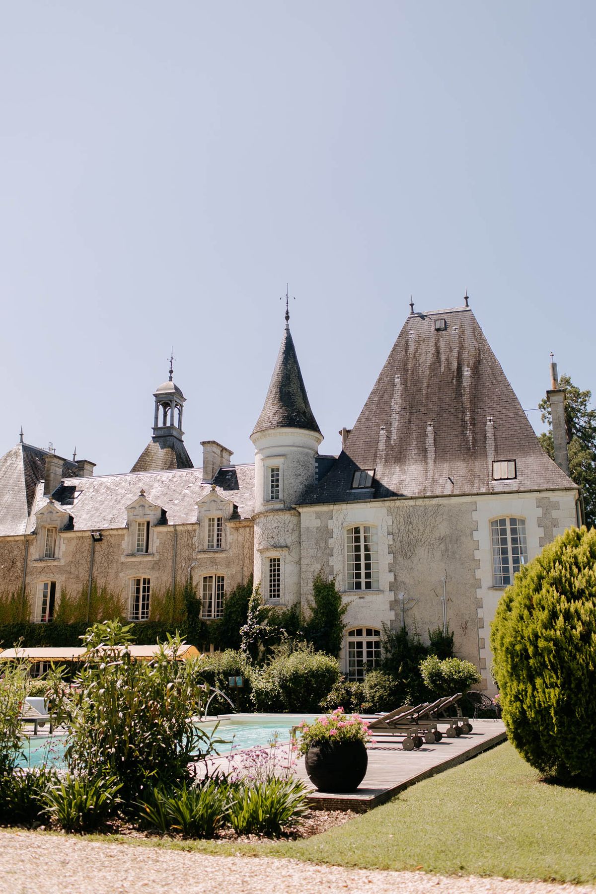 A wide exterior shot of a French château featuring classic architecture with grey slate conical turret roofs, a small bell tower, and cream-toned stone facades with tall arched windows. In the foreground, a rectangular outdoor swimming pool is flanked by a wooden deck with dark sun loungers, a large black spherical planter holding pink flowering plants, and neatly trimmed topiary shrubs. No people are visible in the image. The shot is taken in bright daylight and captures the full rear or side elevation of the building along with its landscaped garden grounds. Potential venue feature image.