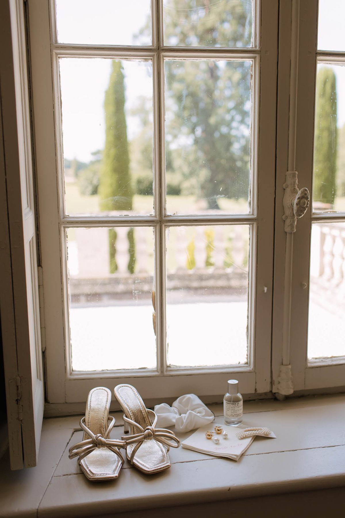 Bridal accessories on windowsill gold knotted sandals Le Labo fragrance pearl earrings and silk scrunchie with garden view