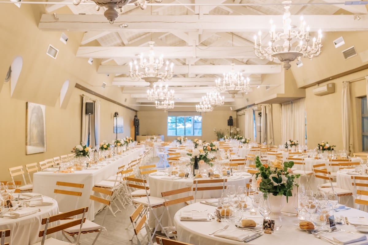 A wide shot of an indoor wedding reception dining room set up and ready for guests, with no people present. The room features warm cream-painted walls, white-painted exposed timber roof trusses, and a row of four white ornate candelabra-style chandeliers running the length of the ceiling. The layout includes a mix of long rectangular tables and round tables, all covered in white linen, surrounded by white metal folding chairs with natural wood slat backs and seats. Centerpieces consist of low arrangements of blush and peach roses with green eucalyptus leaves in white vessels, with small votive candles and glassware completing each place setting. The decor palette is white, blush, and peach with natural wood accents, giving a classic French countryside style. Potential venue feature image.