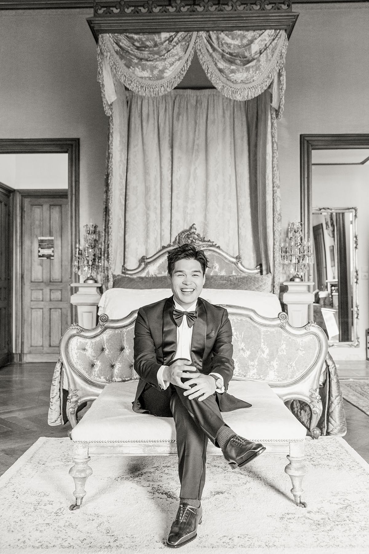 Black and white portrait of groom laughing on ornate canopied bed in chateau suite wearing tuxedo