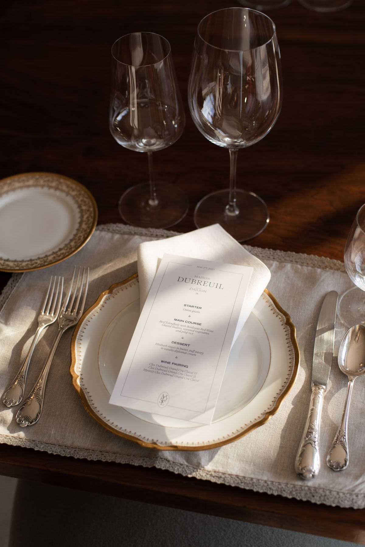 Reception place setting with gold-rimmed china plate, printed menu card, crystal glasses, and ornate silver flatware
