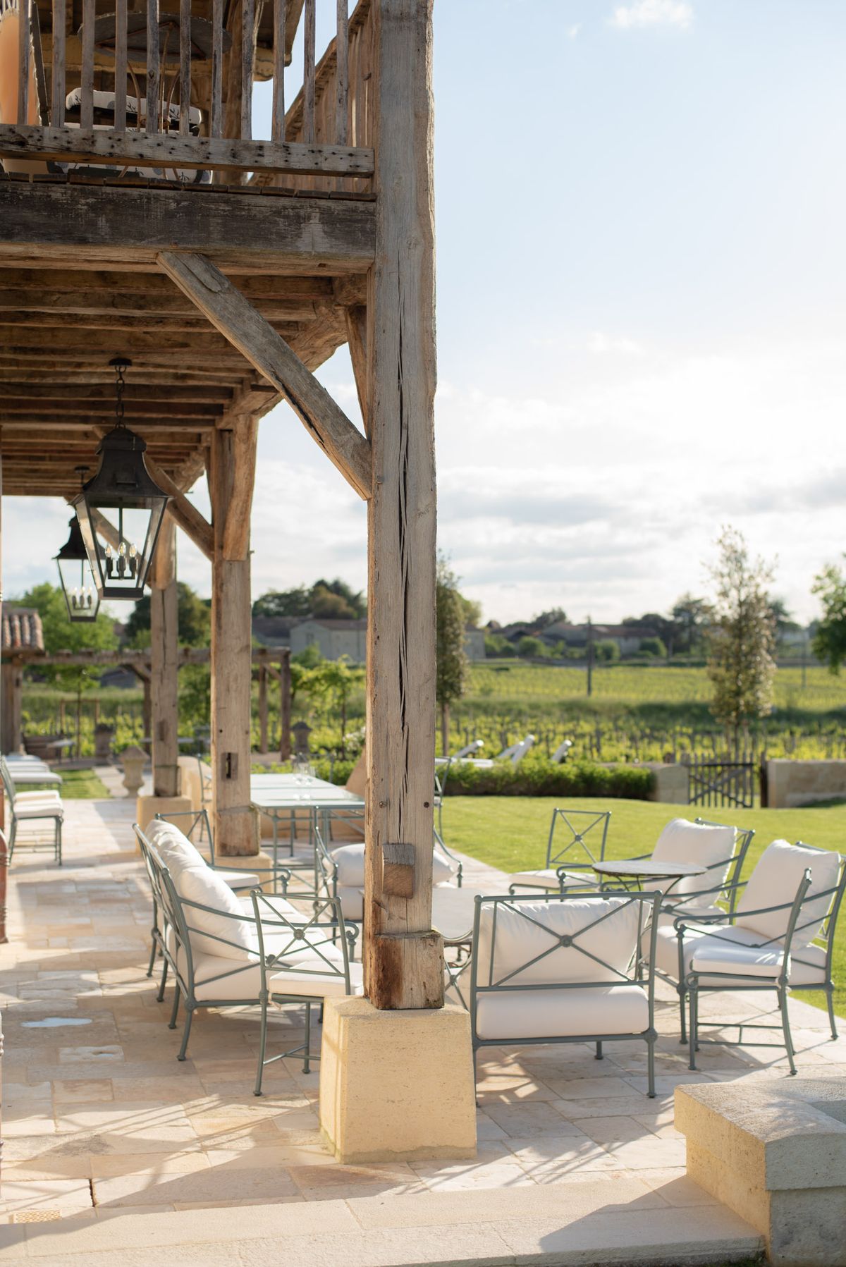 An outdoor terrace at a vineyard property, photographed with no people present. The covered terrace features exposed aged wooden beam framework with a balcony above, and two black hanging lanterns with candle-style bulbs suspended from the ceiling. The seating area is furnished with sage green wrought iron armchairs and a matching small side table, all with white cushions, arranged on a pale limestone-tiled floor. Beyond the covered section, the terrace opens to a manicured lawn with views over rows of vines stretching into the distance, with additional sun loungers visible further along the terrace. The overall style is rustic-refined, combining raw timber construction with polished iron garden furniture. Wide establishing shot. Potential venue feature image.