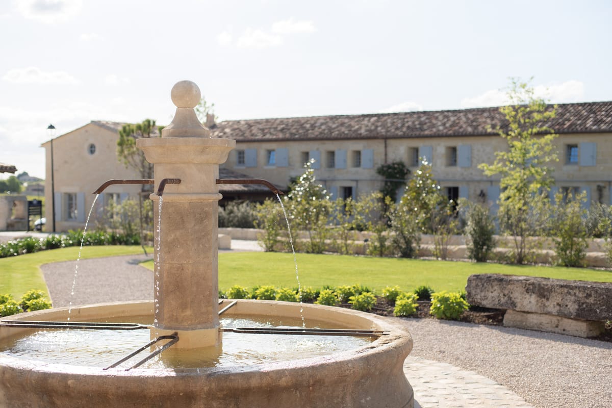 Stone fountain with copper spouts on gravel courtyard before French country estate with blue-grey shutters