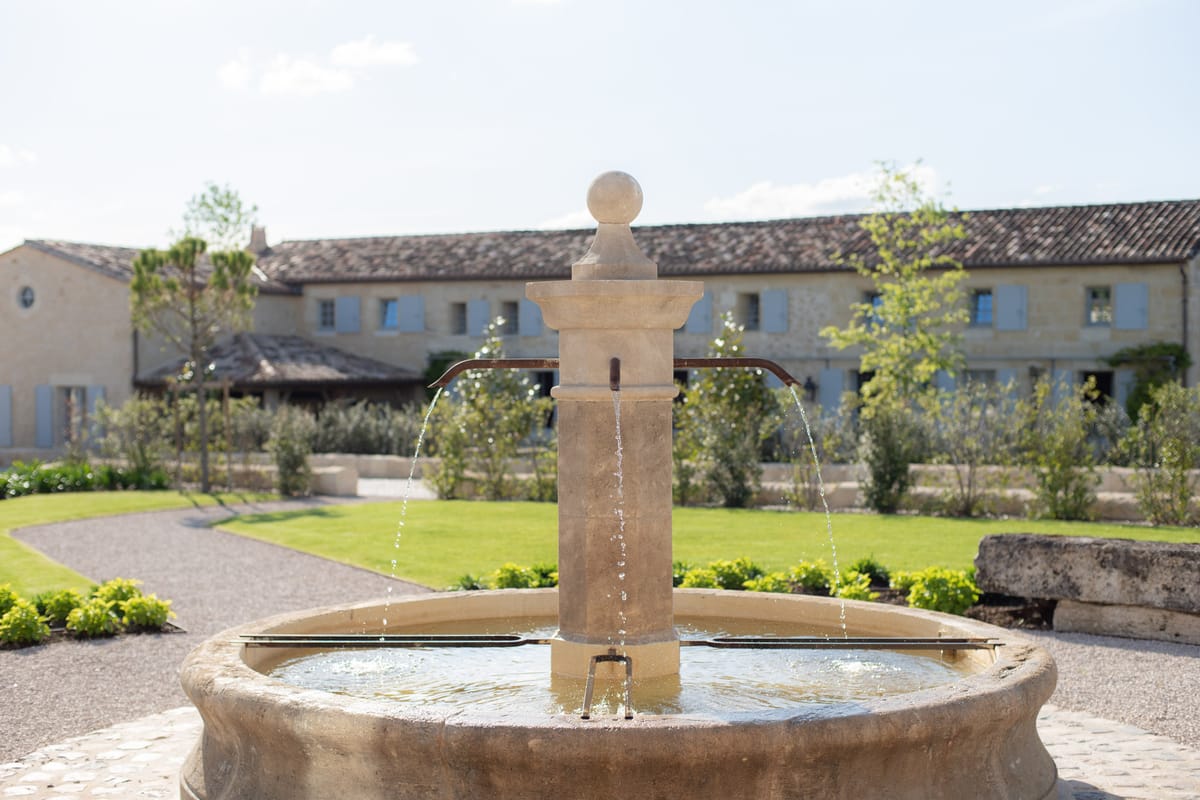 An outdoor shot of a traditional stone fountain in the foreground, with water flowing from four iron spouts into a wide circular basin. Behind it, a formal courtyard garden features a gravel pathway, low boxwood hedging, and young trees arranged in a structured layout. In the background stands a large Provençal-style stone building with terracotta roof tiles, pale blue shutters, and a long symmetrical facade consistent with a French farmhouse or domaine. No people are visible. The composition is a medium close-up on the fountain with the venue building softly out of focus in the background. Potential venue feature image.