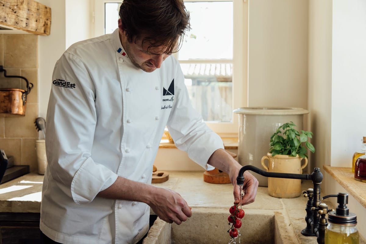 Chef rinsing cherry tomatoes at carved stone sink in Provencal kitchen with copper cookware