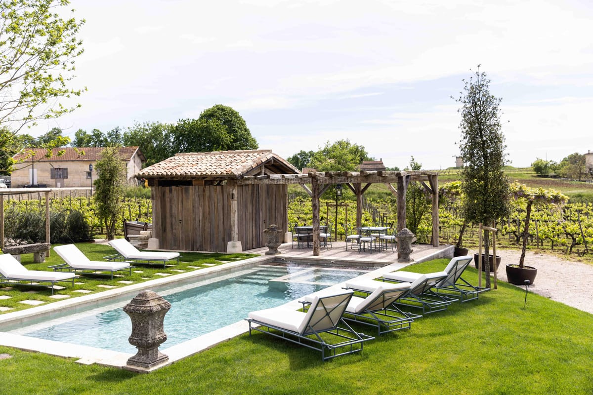 Rectangular pool with white sun loungers and rustic wooden cabana surrounded by vineyard rows at a Bordeaux property