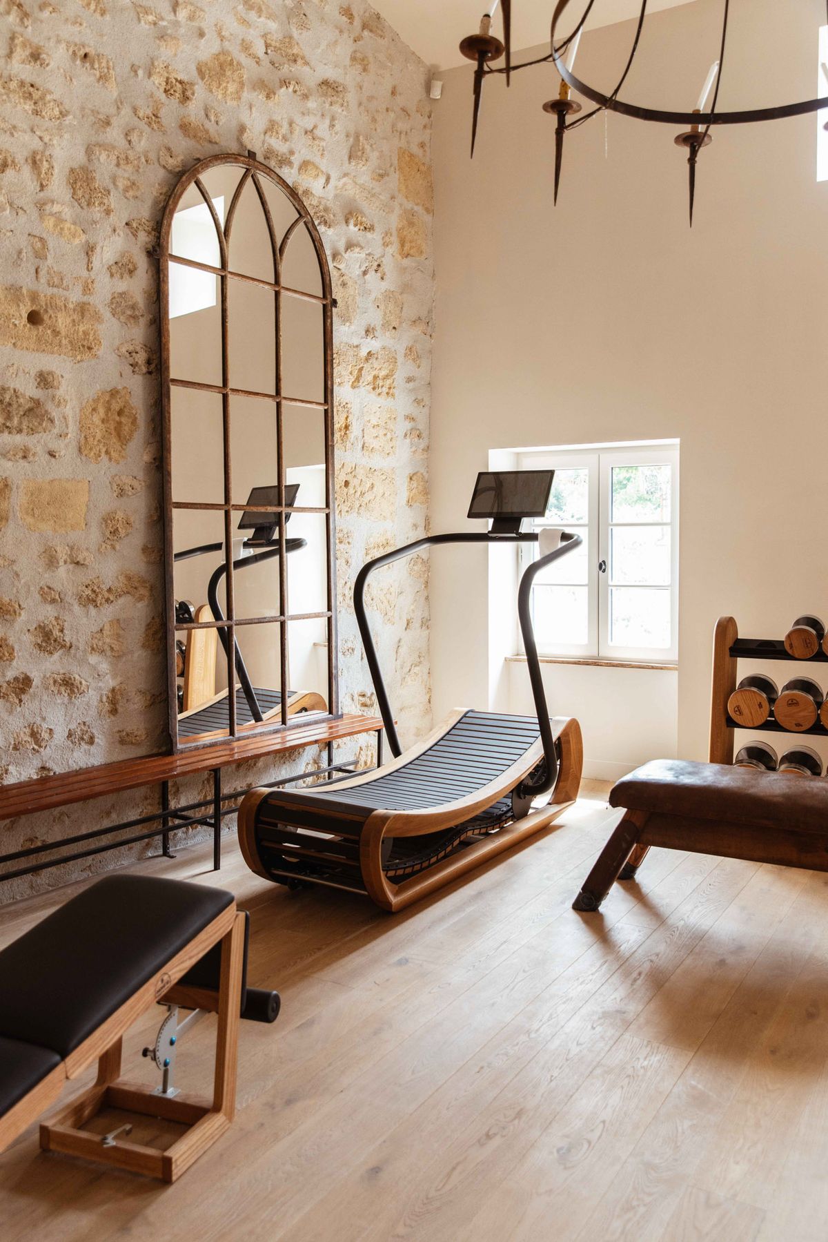 This image does not depict a wedding photograph. It shows the interior of a fitness or wellness room, likely within a French château or manor property. The room features a curved wooden-framed treadmill, a black upholstered weight bench, a wooden dumbbell rack, and a large arched mirror with a dark iron frame leaning against an exposed limestone stone wall. The space has wide-plank light wood flooring, a wrought iron chandelier overhead, and a white-framed French window letting in natural light. The overall aesthetic blends historic architectural features with high-end fitness equipment. Potential venue feature image.