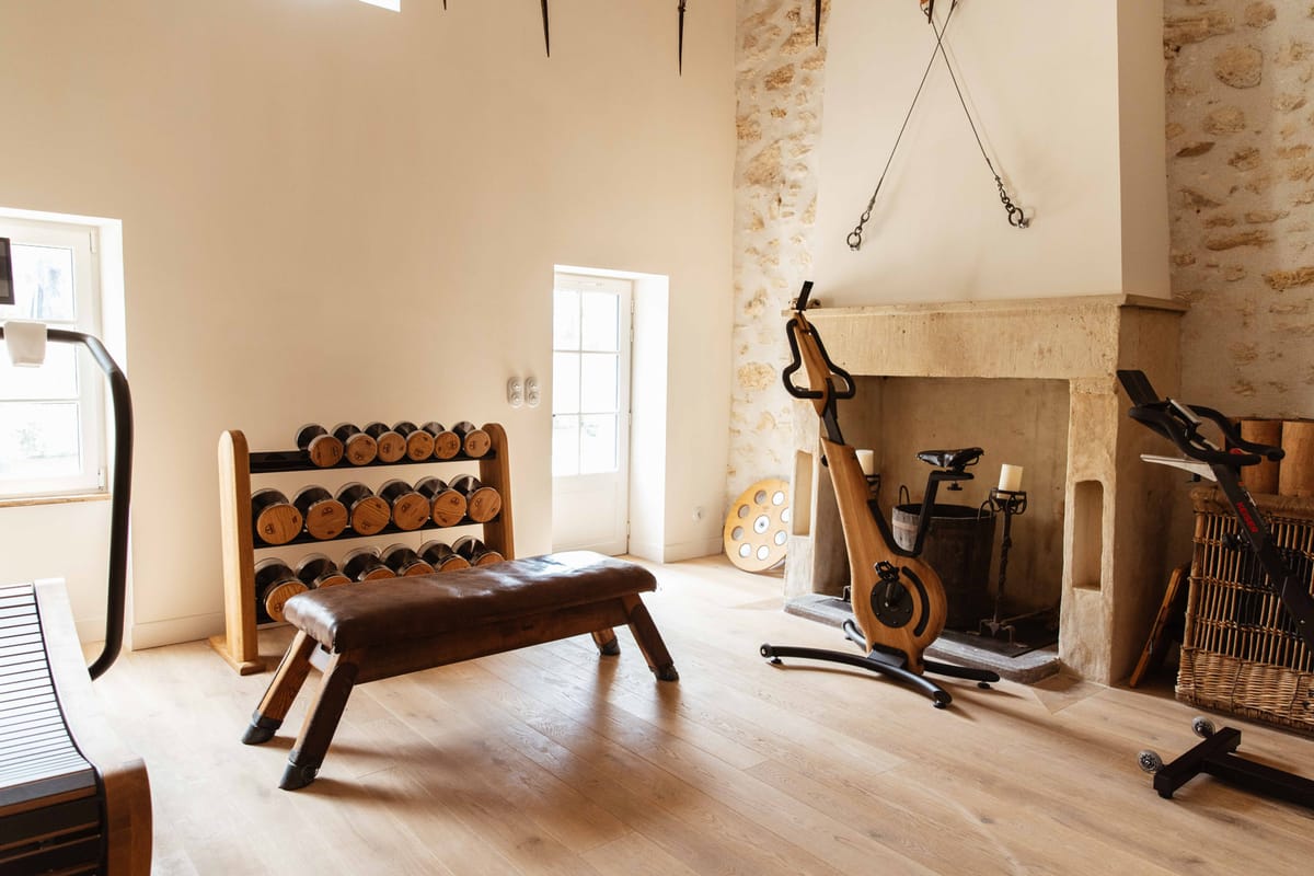 This image shows an interior fitness room within what appears to be a French château or manor property. The space features high ceilings with white-plastered walls, a large stone fireplace surround, and light oak herringbone-style flooring. The room is equipped with a wooden dumbbell rack holding a full set of matching wood-and-metal dumbbells, a brown leather gymnastics pommel bench, a wood-frame stationary cycling bike, a cable pulley system mounted near the ceiling, and additional cardio equipment partially visible at the edges of the frame. The overall aesthetic blends contemporary fitness equipment with warm wood tones against the historic stone architectural details. Wide interior shot with natural light entering through two windows and a glass-paned door. Potential venue feature image.