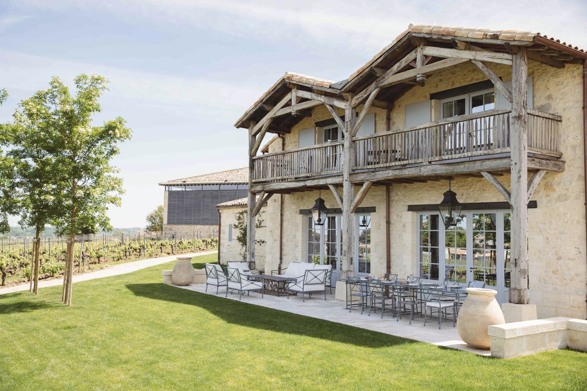 Wide exterior shot of a two-story French stone and timber-frame building set within a vineyard property. The facade features weathered wooden beam framing, a wraparound upper balcony with wooden railings, black iron pendant lanterns, and tall French doors opening onto a stone-paved terrace. The terrace is furnished with wrought-iron garden chairs and sofas with white cushions arranged around a long dining table, suggesting an outdoor reception or cocktail setup. Rows of grapevines extend behind the building, with a secondary stone outbuilding visible in the background. The overall aesthetic is rustic French countryside with a refined, understated palette of warm stone, weathered grey wood, and white upholstery. No people are present. Potential venue feature image.