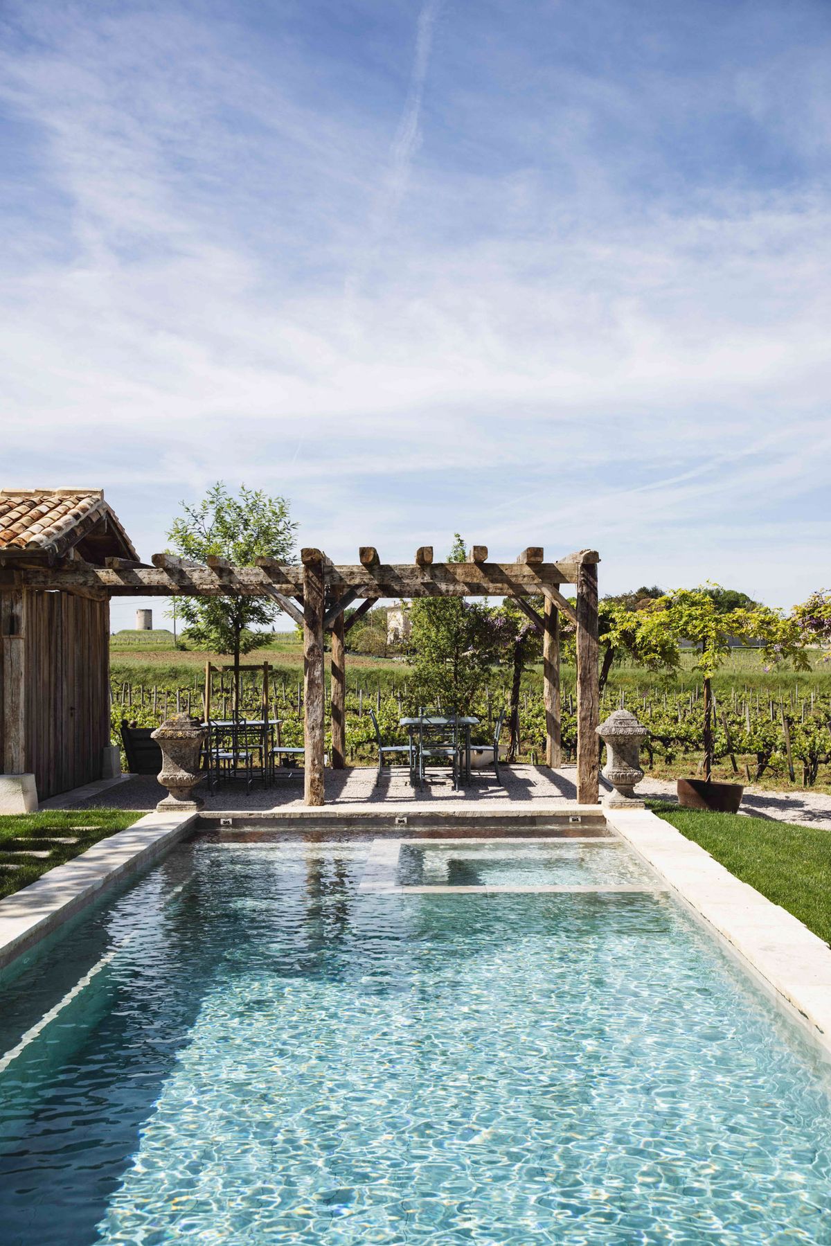 An outdoor venue shot showing a rectangular swimming pool with a pale stone surround, photographed from pool level looking toward a rustic wooden pergola structure. Beneath the pergola sit two small dining tables with metal chairs, flanked by a pair of large stone urn planters. A weathered timber outbuilding with a terracotta-tiled roof is visible to the left. The property borders a vineyard that extends across the background, with rows of vines stretching to the horizon. The overall aesthetic is rustic French countryside with natural wood, aged stone, and iron furniture. Wide establishing shot taken in daylight. Potential venue feature image.