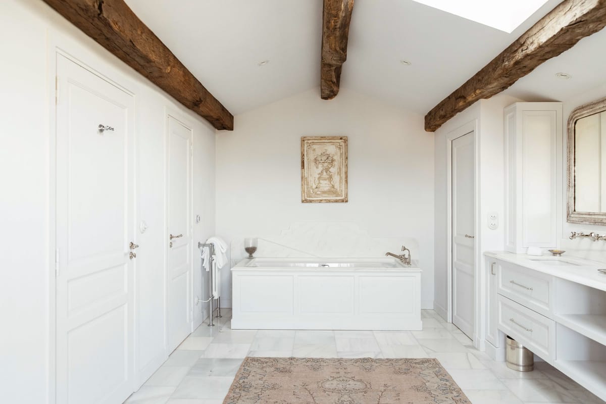A wide-shot interior image of a venue bathroom, showing no people or wedding activity. The room features an all-white palette with a built-in white panelled bathtub centered against the back wall, white cabinetry with silver hardware, a white marble or stone vanity counter, and a skylight overhead. Exposed dark wood ceiling beams run the length of the vaulted ceiling, contrasting with the white walls and marble tile floor. A faded dusty rose and taupe vintage-style area rug is placed on the floor in front of the tub, and an antique-style decorative panel in muted gold and cream tones hangs on the wall above the bath. A silver towel rail with white folded towels and a silver-framed mirror on the right wall complete the decor. The overall style mixes rustic French farmhouse elements with classic, clean interior design. Potential venue feature image.