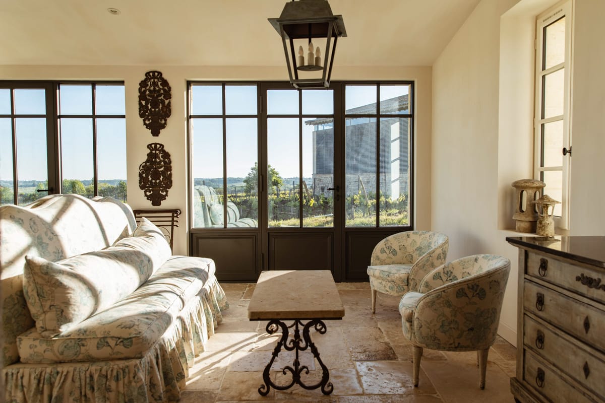 An interior sitting room at what appears to be a French countryside venue, likely a château or domaine, shown with no people present. The room features a cream and soft blue-green floral skirted sofa, two matching floral barrel chairs, and a stone-topped wrought iron coffee table arranged on large-format limestone tile flooring. A dark iron pendant lantern hangs from the ceiling, and two ornate dark iron decorative wall medallions are mounted between tall steel-framed French doors and windows that look out onto a vineyard and outbuilding. A distressed grey wooden chest of drawers with iron ring pulls sits to the right, topped with a weathered stone decorative object. The overall interior palette is warm cream, aged stone, and muted blue-green, with a classic French country styling theme. Wide interior shot. Potential venue feature image.