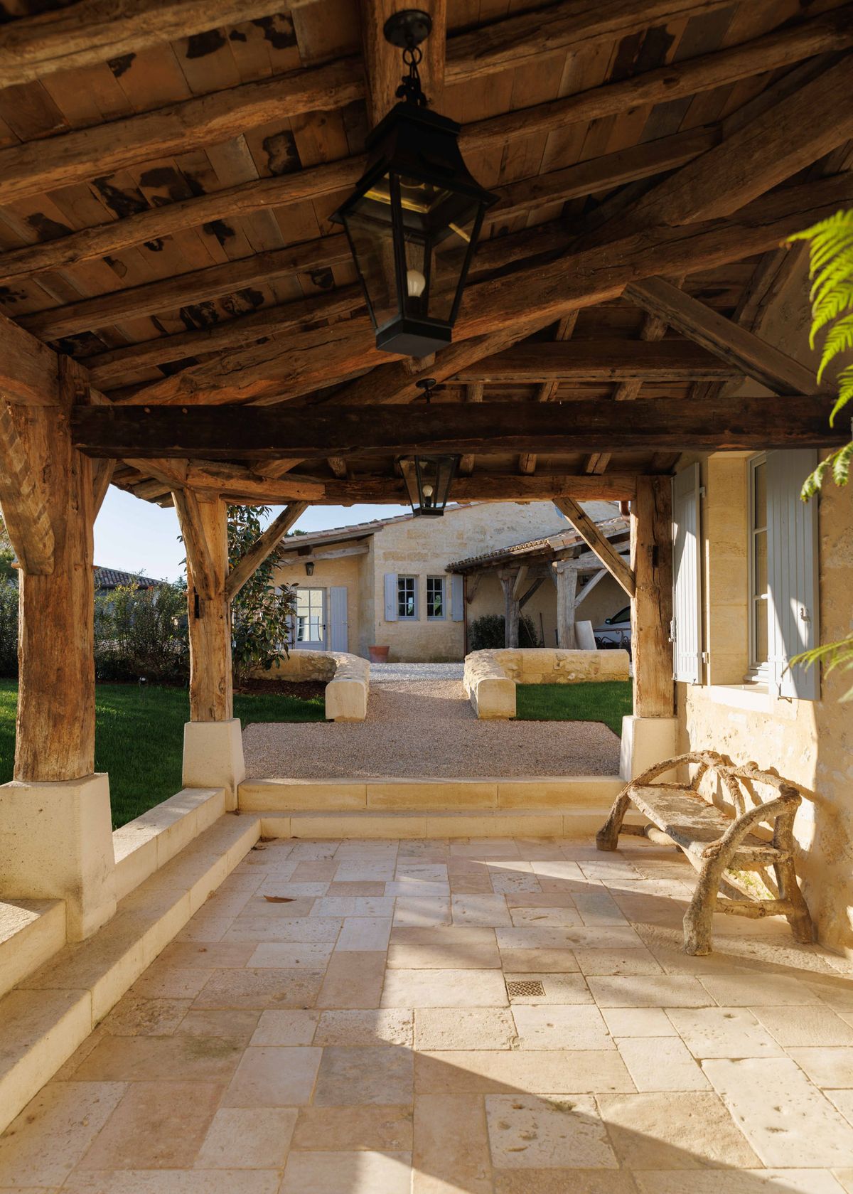 A wide-angle architectural shot of a covered outdoor terrace at a French rural property, with no people present. The structure features heavy reclaimed timber beams and posts set on cream-colored stone bases, with a planked wooden ceiling from which two black iron lanterns hang. The pale limestone tile floor leads out to a gravel pathway flanked by low stone benches and a manicured lawn, with a secondary stone farmhouse building visible in the background featuring grey-blue shutters. A rustic twig-and-branch bench sits to the right of the frame. The styling is distinctly rustic French countryside, with warm golden afternoon light casting sharp shadows across the terrace. No wedding-specific decor is visible. Potential venue feature image.