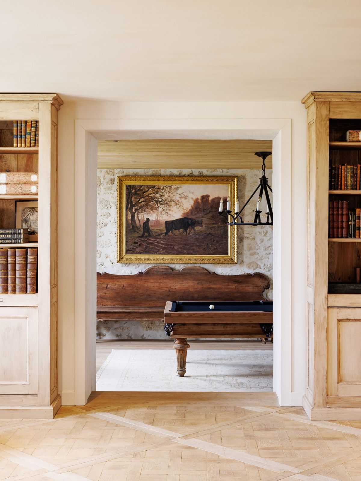 Interior venue shot showing two adjoining rooms viewed through a wide doorway arch. The foreground room features light wood parquet flooring in a chevron pattern and two built-in natural wood bookcases flanking the doorway, filled with antique leather-bound books. Through the doorway, a billiards table with a dark navy felt surface and ornate carved dark walnut frame sits on a pale stone floor. Behind it, an antique carved wooden bench with a scalloped back is positioned against an exposed limestone or white stone wall. A large oil painting in a gilded gold frame depicting a pastoral farming scene with cattle hangs on the wall, and a black wrought-iron chandelier with candle-style lights is suspended from a wood-beamed ceiling. The overall interior style is French country classic, with a warm, neutral palette of natural wood tones, cream, and warm brown. Wide shot, no people present. Potential venue feature image.