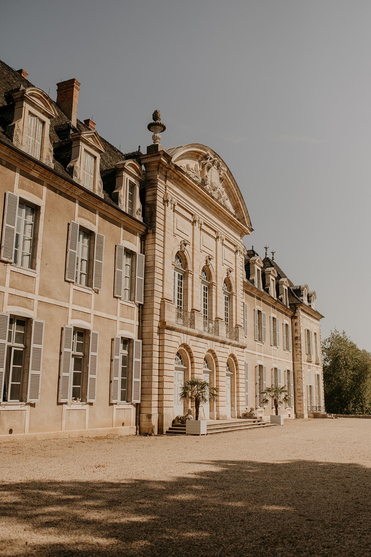 An outdoor wide-angle shot of a large French classical chateau facade, shot from a low angle along the gravel forecourt. The building features cream-toned limestone architecture with a prominent central pavilion topped by a carved pediment with decorative stonework and a stone finial, arched ground-floor doorways, wrought-iron balconettes, and white-shuttered windows across multiple stories with a mansard slate roof. Two potted palm trees flank the central entrance steps, and the gravel courtyard extends across the foreground. No people are visible in the frame. Potential venue feature image.