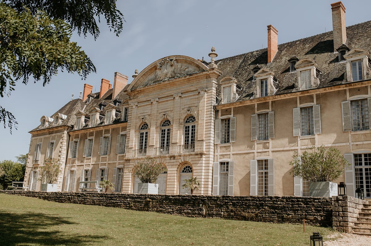 Golden stone chateau with slate mansard roof, arched windows, white shutters, and planter-lined terrace