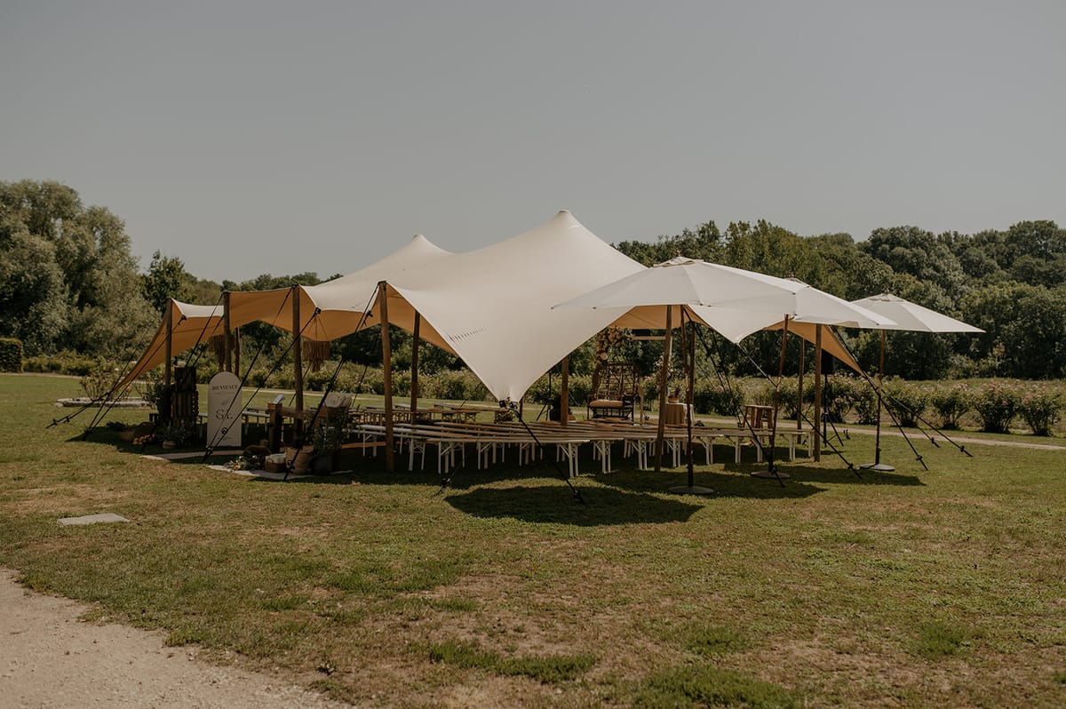 A wide shot of an outdoor wedding reception setup on a large lawn, featuring a freeform stretch tent in cream and warm ochre tones supported by wooden poles, with additional large parasols extending the covered area. Beneath the tent structure, long white trestle tables with benches are arranged in rows on a raised wooden deck platform, styled in a bohemian-rustic aesthetic. A floral arch in warm tones is visible at the back of the setup near what appears to be a sweetheart table or focal seating area, and a welcome sign is positioned at the entrance. No people are visible in the image, suggesting this was taken before guests arrived. Potential venue feature image.