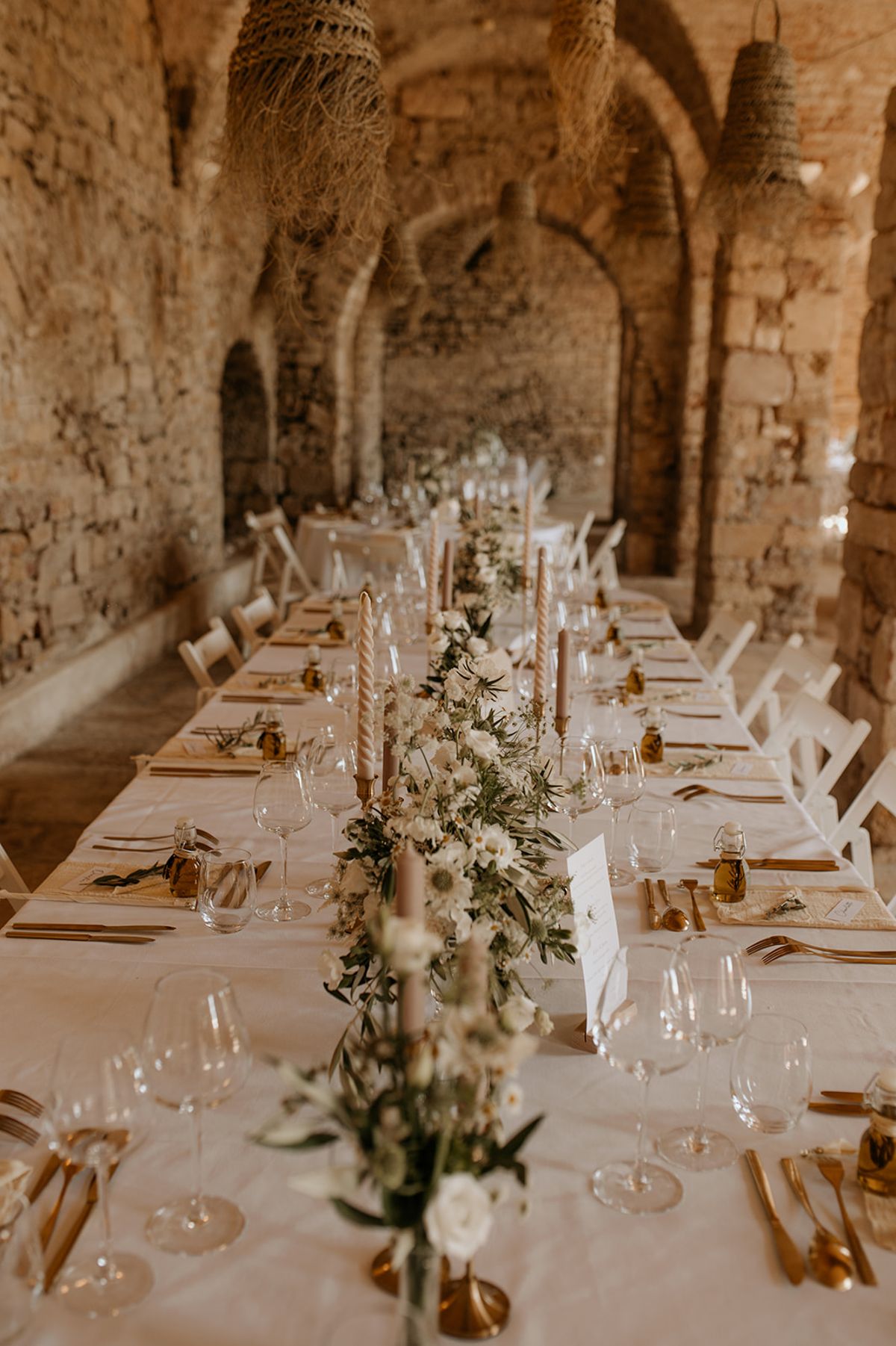 Long table with white rose and olive runner, terracotta tapers, and rattan pendants in vaulted stone cellar