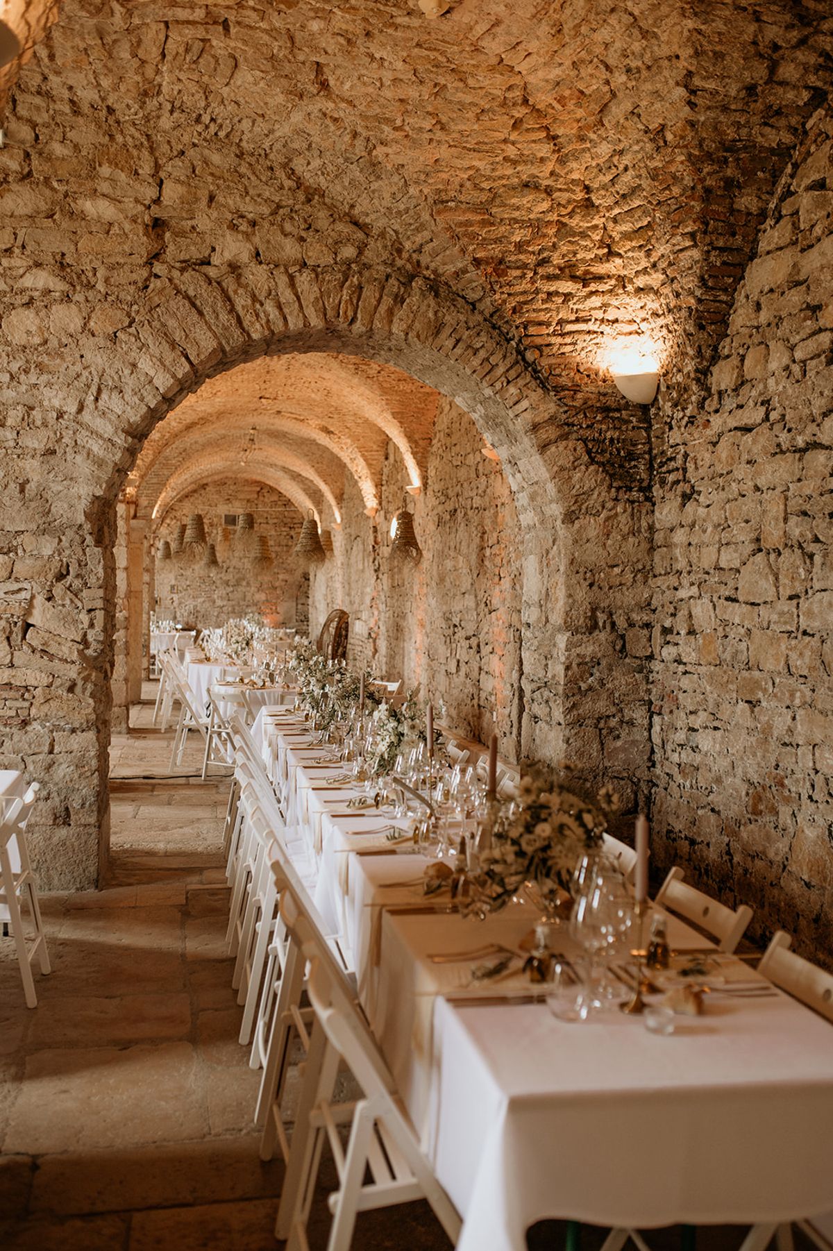 A wedding reception setup photographed in a vaulted stone room, likely a medieval cellar or abbey, with a series of rounded arched passageways receding into the background. Long banquet tables dressed in white linen run along the length of the space, flanked by white folding chairs. The tablescape features a low, continuous floral runner of white and cream blooms — appearing to include roses and gypsophila — with greenery, slender taper candles in nude tones, gold cutlery, and crystal glassware. Warm wall-mounted uplighting illuminates the honey-toned stone arches. The composition is a wide perspective shot taken from one end of the room, emphasizing the repeating arches and the length of the table. The overall decor palette is white, cream, and gold against the raw stone architecture. Potential venue feature image.