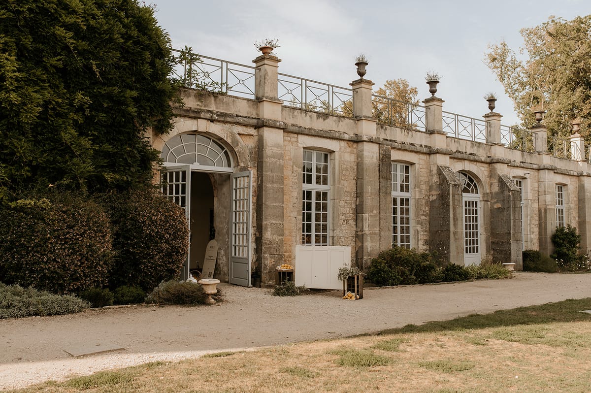An exterior wide shot of a French château outbuilding or orangery, constructed in pale honey-colored stone with a flat roofline featuring decorative iron railings and stone urns with plantings along the top. The façade includes tall arched windows with white-painted wooden frames, a central arched doorway with fanlight detail standing open, and a gravel forecourt in the foreground. Near the entrance, wooden crates filled with what appear to be yellow citrus fruits or melons are placed alongside a white painted cabinet and a decorative wooden board, suggesting event setup or a welcome display for a wedding. No people are visible in the frame. Potential venue feature image.